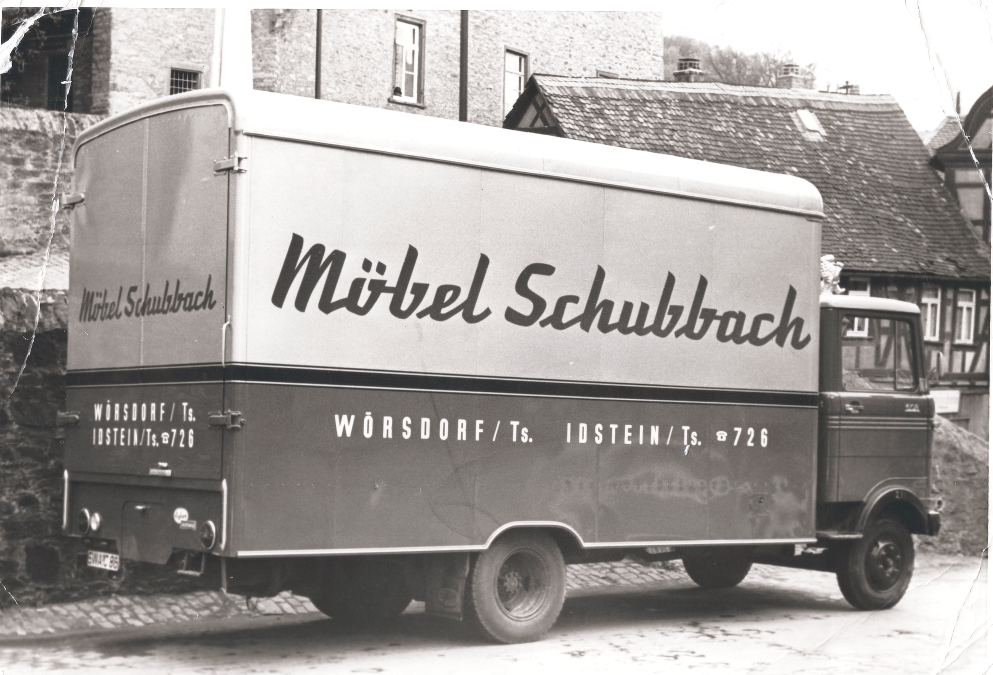 Ein nostalgischer Möbeltransporter mit der Aufschrift "Möbel Schubbach", parkend vor historischen Gebäuden.
