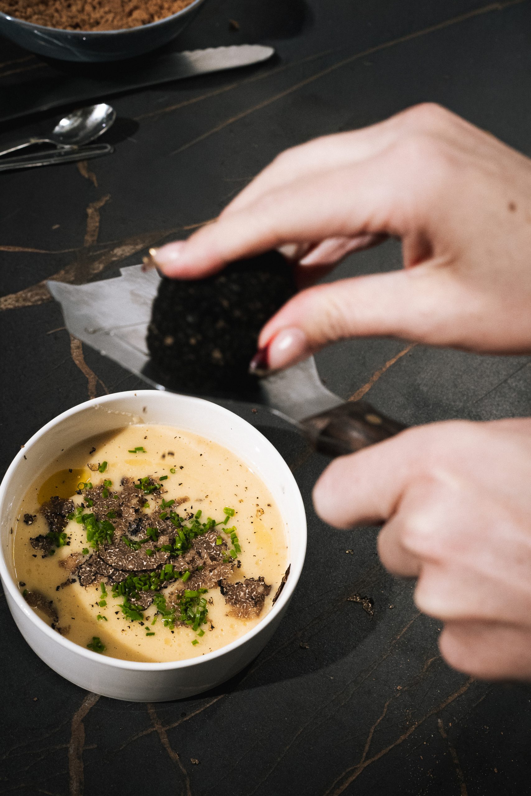 Eine Hand schneidet schwarzen Trüffel über eine cremige Suppe, garniert mit Kräutern und Trüffelscheiben.
